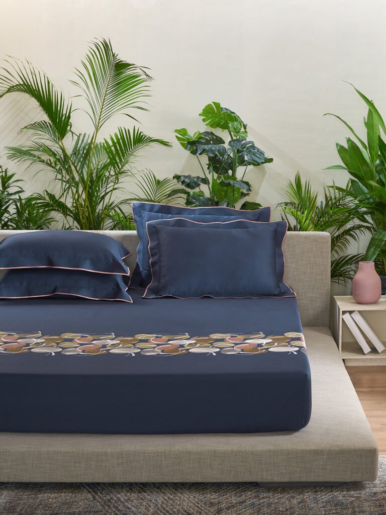 A bed with blue Welspun Bedsheets and pillows placed in front of a plant.